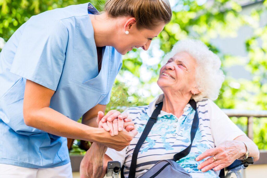 Pflegekraft begrüßt Seniorin im Rollstuhl im Garten.