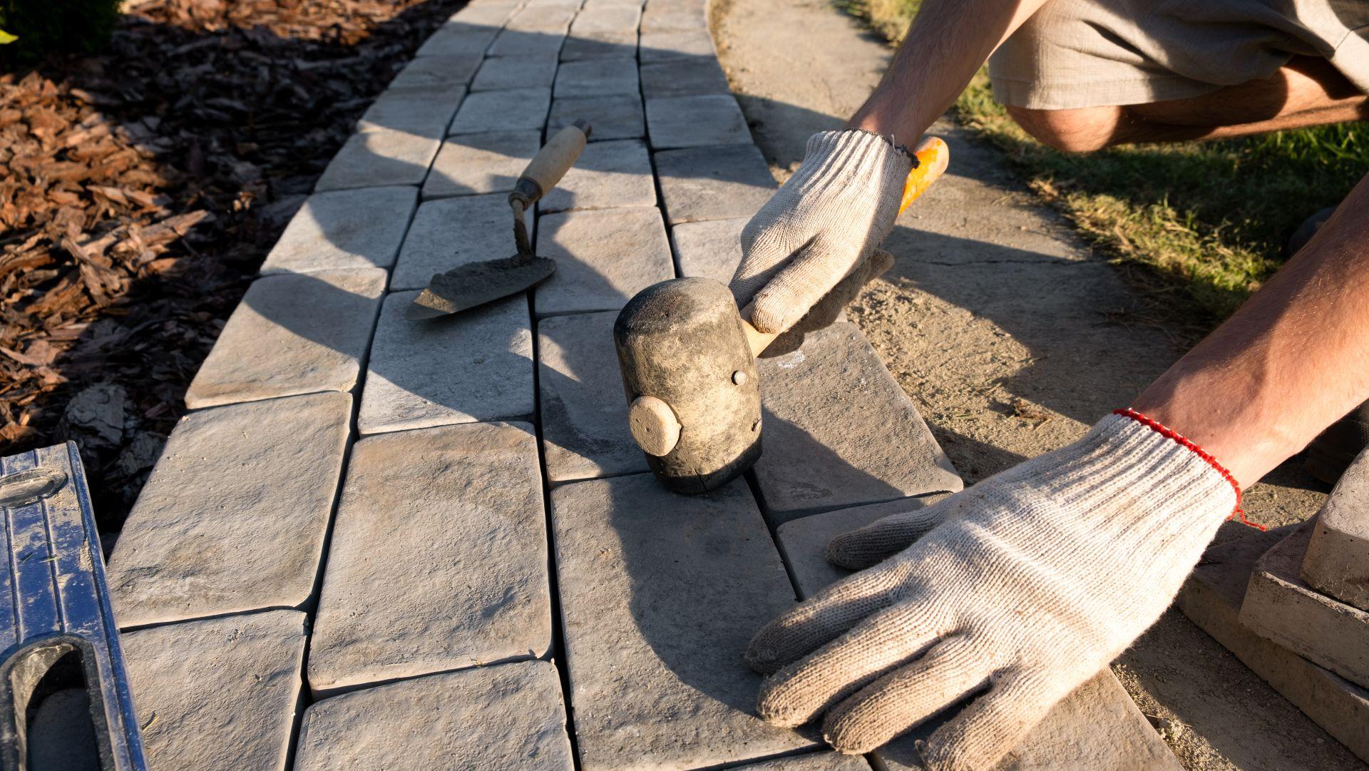 Ein Handwerker mit Arbeitshandschuhen richtet Kopfsteinpflaster sorgfältig in einer Sandbettung aus. Neben ihm liegen Werkzeuge wie Wasserwaage und Kelle. Das Bild zeigt den präzisen Arbeitsprozess beim Pflasterverlegen und betont handwerkliche Qualität und Stabilität.